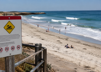 Beach warning sign in the foreground overlooking a sunny shoreline with waves and scattered beachgoers, used as an illustrative image for an L.A. County ocean water quality warning during a Southern California heat wave.