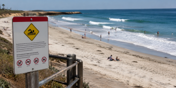 Beach warning sign in the foreground overlooking a sunny shoreline with waves and scattered beachgoers, used as an illustrative image for an L.A. County ocean water quality warning during a Southern California heat wave.