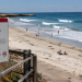 Beach warning sign in the foreground overlooking a sunny shoreline with waves and scattered beachgoers, used as an illustrative image for an L.A. County ocean water quality warning during a Southern California heat wave.