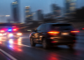 Blurred image of a dark SUV on a city roadway with red and blue emergency lights behind it, used as an illustrative cover for a high-speed pursuit involving a road rage suspect across Los Angeles County.