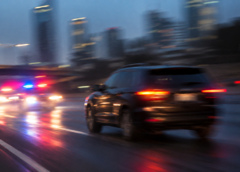 Blurred image of a dark SUV on a city roadway with red and blue emergency lights behind it, used as an illustrative cover for a high-speed pursuit involving a road rage suspect across Los Angeles County.