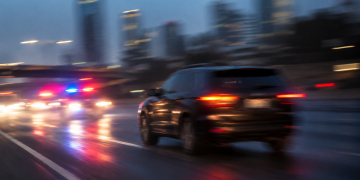 Blurred image of a dark SUV on a city roadway with red and blue emergency lights behind it, used as an illustrative cover for a high-speed pursuit involving a road rage suspect across Los Angeles County.
