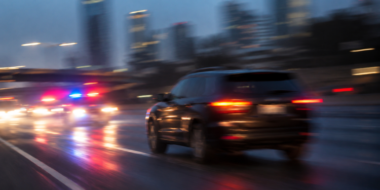 Blurred image of a dark SUV on a city roadway with red and blue emergency lights behind it, used as an illustrative cover for a high-speed pursuit involving a road rage suspect across Los Angeles County.