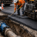 Close-up of construction crews repairing an excavated roadway with an exposed water pipe, fresh asphalt and paving equipment, used as an illustrative image for the West Hollywood Sunset Boulevard sinkhole repair.