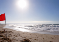 Red warning flag on a sunny beach with rough waves and bright heat haze, used as an illustrative image for dangerous surf and persistent heat across Southern California.