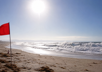 Red warning flag on a sunny beach with rough waves and bright heat haze, used as an illustrative image for dangerous surf and persistent heat across Southern California.