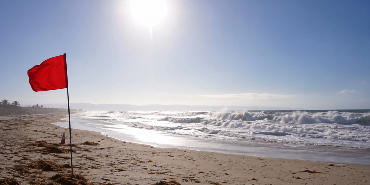 Red warning flag on a sunny beach with rough waves and bright heat haze, used as an illustrative image for dangerous surf and persistent heat across Southern California.