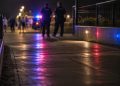 Blurred police officers and distant pedestrians near a coastal walkway at night, with red and blue emergency lights reflecting on the pavement.