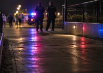 Blurred police officers and distant pedestrians near a coastal walkway at night, with red and blue emergency lights reflecting on the pavement.