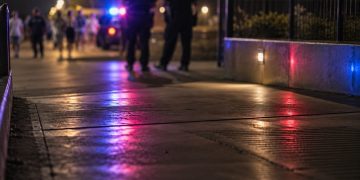 Blurred police officers and distant pedestrians near a coastal walkway at night, with red and blue emergency lights reflecting on the pavement.