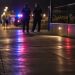 Blurred police officers and distant pedestrians near a coastal walkway at night, with red and blue emergency lights reflecting on the pavement.