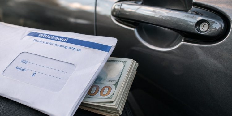 Close-up of a bank withdrawal envelope beside a car door handle and folded cash.
