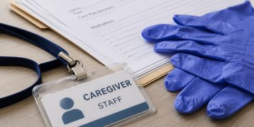 Close-up of a caregiver identification badge, disposable gloves and paperwork on a neutral desk.