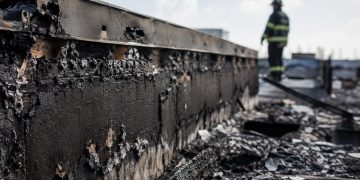 Close-up of a charred commercial roof edge and burned debris, with a blurred firefighter monitoring the damaged structure in the background.