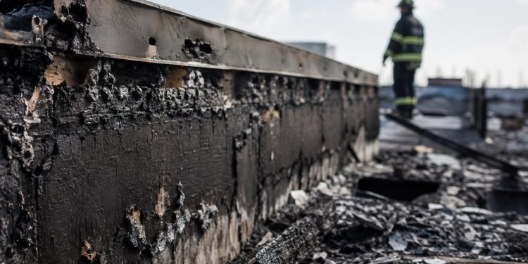 Close-up of a charred commercial roof edge and burned debris, with a blurred firefighter monitoring the damaged structure in the background.