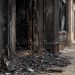 Close-up of a charred storefront entrance and burned debris after a commercial fire, with a blurred firefighter standing farther down the sidewalk.