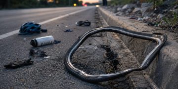 Close-up of a damaged bicycle wheel and scattered cycling gear beside a curb, with a blurred investigator standing farther down the road.