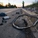 Close-up of a damaged bicycle wheel and scattered cycling gear beside a curb, with a blurred investigator standing farther down the road.