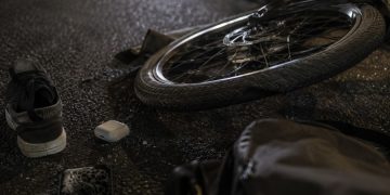 Close-up of a damaged e-bike wheel and scattered personal belongings on dark pavement.