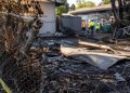 Close-up of a damaged fence and burned debris outside a vacant residential property, with blurred city workers standing farther back.