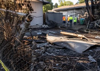 Close-up of a damaged fence and burned debris outside a vacant residential property, with blurred city workers standing farther back.