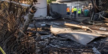 Close-up of a damaged fence and burned debris outside a vacant residential property, with blurred city workers standing farther back.