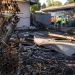 Close-up of a damaged fence and burned debris outside a vacant residential property, with blurred city workers standing farther back.