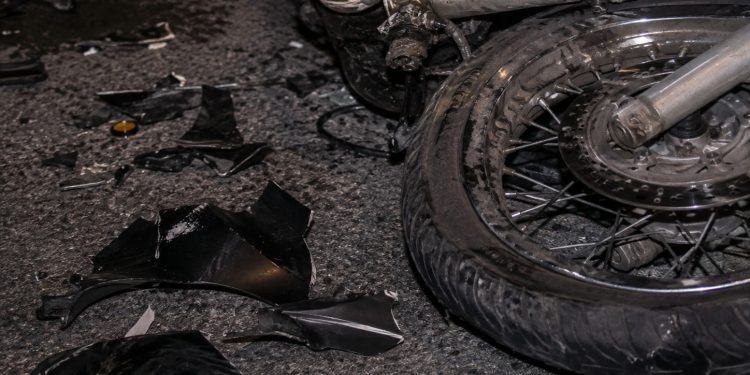 Close-up of a damaged motorcycle wheel and broken plastic pieces scattered across dark pavement.