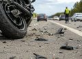 Close-up of a damaged motorcycle wheel and scattered roadway debris on freeway pavement, with blurred traffic and a responder in the distance.