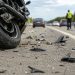 Close-up of a damaged motorcycle wheel and scattered roadway debris on freeway pavement, with blurred traffic and a responder in the distance.