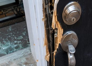 Close-up of a damaged residential door lock and splintered frame beside scattered glass.