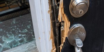 Close-up of a damaged residential door lock and splintered frame beside scattered glass.
