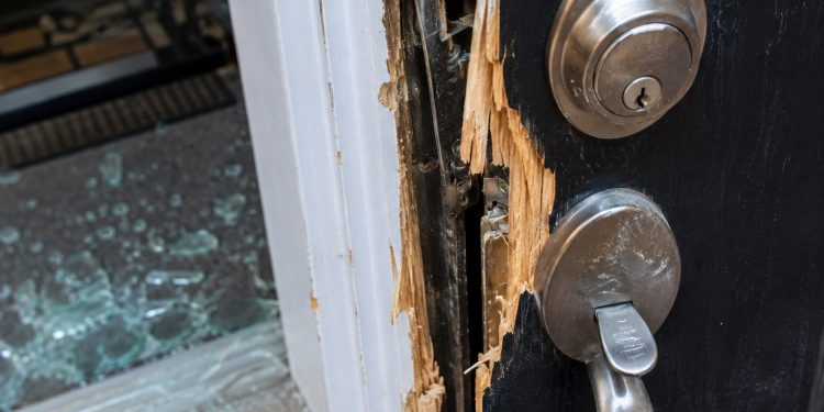 Close-up of a damaged residential door lock and splintered frame beside scattered glass.