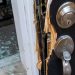 Close-up of a damaged residential door lock and splintered frame beside scattered glass.