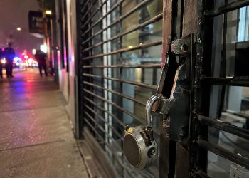 Close-up of a damaged security gate and broken storefront lock, with blurred people and police lights farther down the street.