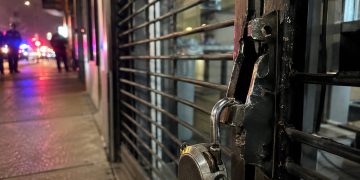 Close-up of a damaged security gate and broken storefront lock, with blurred people and police lights farther down the street.
