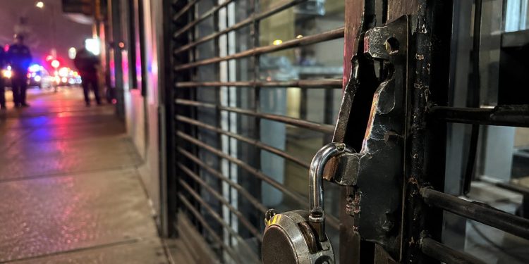Close-up of a damaged security gate and broken storefront lock, with blurred people and police lights farther down the street.