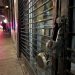 Close-up of a damaged security gate and broken storefront lock, with blurred people and police lights farther down the street.