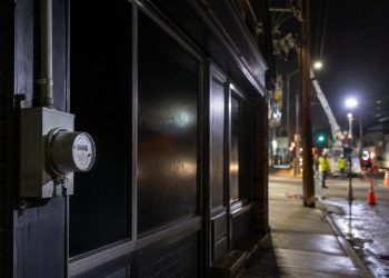 Close-up of a darkened storefront and utility meter at night, with blurred repair crews and work lights farther down the street.