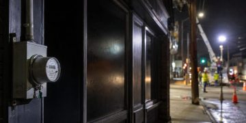 Close-up of a darkened storefront and utility meter at night, with blurred repair crews and work lights farther down the street.