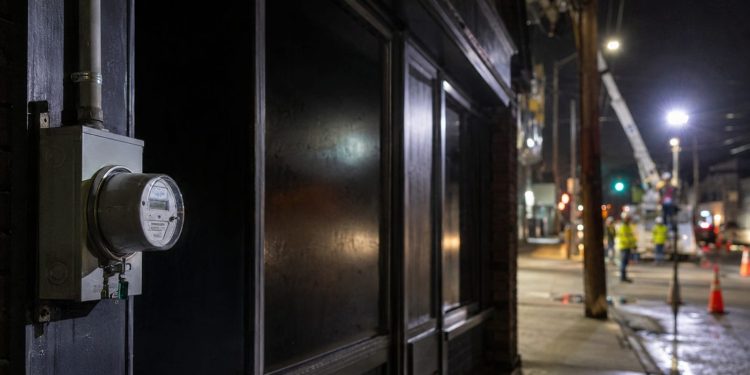 Close-up of a darkened storefront and utility meter at night, with blurred repair crews and work lights farther down the street.