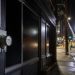 Close-up of a darkened storefront and utility meter at night, with blurred repair crews and work lights farther down the street.