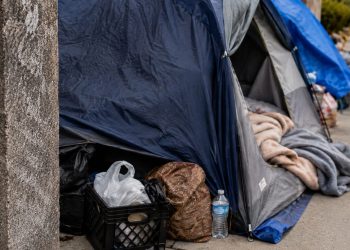 Close-up of a posted sidewalk restriction sign beside tents and personal belongings along an urban walkway.