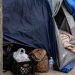 Close-up of a posted sidewalk restriction sign beside tents and personal belongings along an urban walkway.