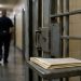 Close-up of a prison cell door and folded legal papers on a metal surface, with a blurred correctional officer walking in the background.