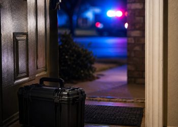 Close-up of a residential doorway with a small bomb-squad equipment case and blurred emergency lights outside.