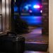 Close-up of a residential doorway with a small bomb-squad equipment case and blurred emergency lights outside.