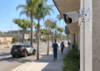 Close-up of a residential security camera overlooking a commercial street, with pedestrians and a heavily blurred patrol vehicle in the background.