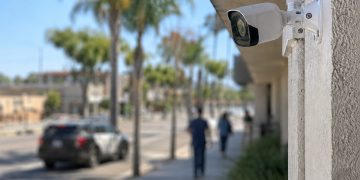 Close-up of a residential security camera overlooking a commercial street, with pedestrians and a heavily blurred patrol vehicle in the background.