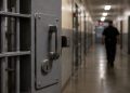 Close-up of a secured prison cell door and dim corridor, with a blurred correctional officer walking in the background.